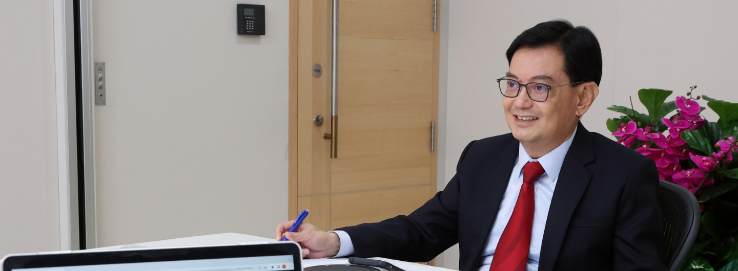 Man in suit with red tie at desk with laptop, holding pen, floral arrangement behind him.